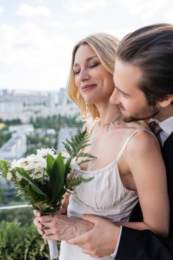 Brunette groom hugging smiling bride with bouquet on terrace 