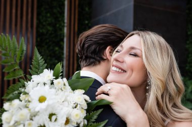 Cheerful bride embracing groom near bouquet outdoors 
