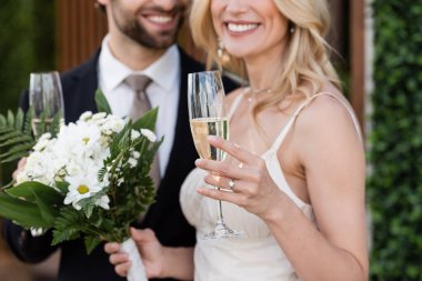 Cropped view of glass of champagne in hand of blurred bride and groom outdoors 