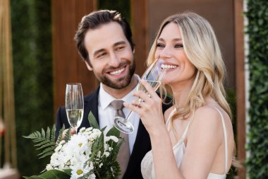 Smiling bride holding glass of champagne near blurred groom and flowers outdoors 