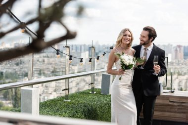 Happy groom holding champagne and looking at blonde bride with bouquet on terrace of restaurant 