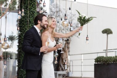 Positive newlyweds with champagne and bouquet standing near light bulbs on terrace of restaurant  clipart