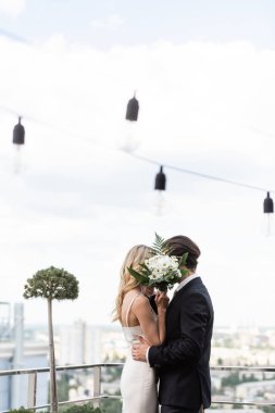 Newlyweds covering faces with bouquet while kissing on terrace 