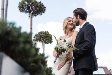 Low angle view of groom looking at smiling bride with flowers on terrace 