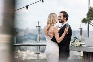 Positive groom hugging bride with glass of champagne on terrace of restaurant 
