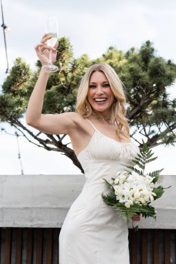 Cheerful bride in wedding dress holding champagne and flowers on terrace 