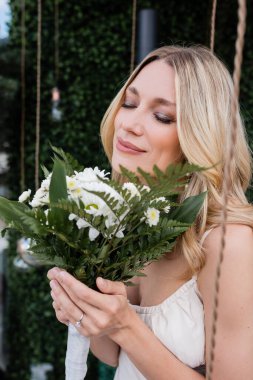 Blonde bride with closed eyes smelling bouquet on terrace 