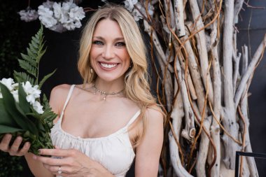 Cheerful bride with flowers looking at camera near decorative twigs on terrace 