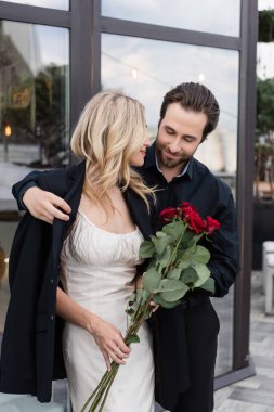 Brunette man wearing jacket on girlfriend with red roses on terrace 