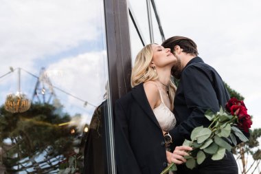 Young man kissing girlfriend with red roses on terrace 