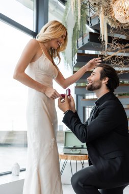 Side view of man holding box with engagement ring near cheerful girlfriend in restaurant 