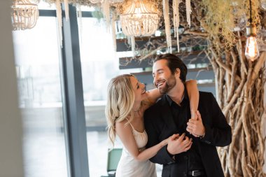 Cheerful woman in dress holding hands of boyfriend under chandelier in restaurant 