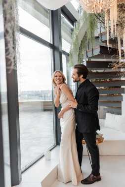 Smiling man looking at elegant girlfriend in dress near window in restaurant 