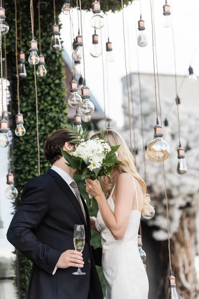 Newlyweds Bouquet Champagne Covering Faces While Kissing Light Bulbs Outdoors — Stock Photo, Image