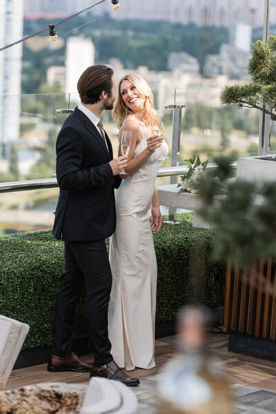 elegant groom holding champagne near smiling bride in dress on terrace 