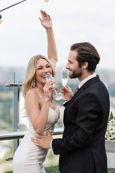 Cheerful blonde bride holding champagne near groom on terrace 