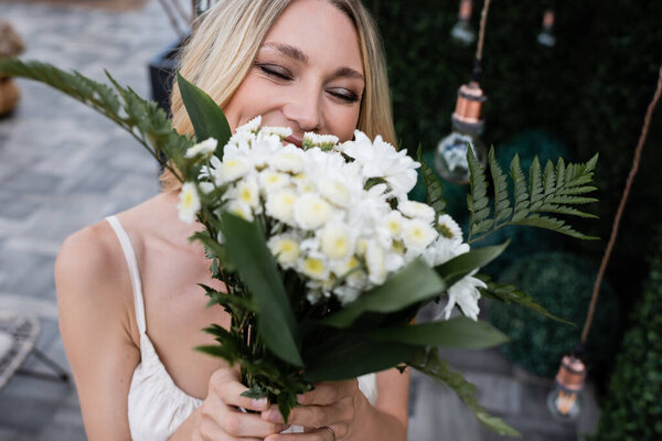 Blonde bride covering face with blurred bouquet on terrace 