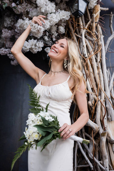 Happy bride in dress holding bouquet near decorative flowers on terrace of restaurant 