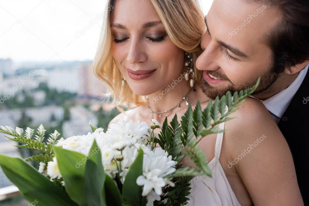 Smiling groom standing near bride with blurred bouquet on terrace