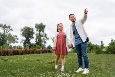 Cheerful asian daughter and dad looking away in summer park  clipart