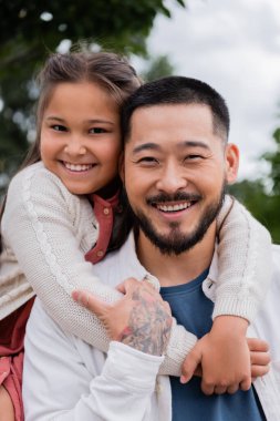 Positive asian girl embracing tattooed father in park 