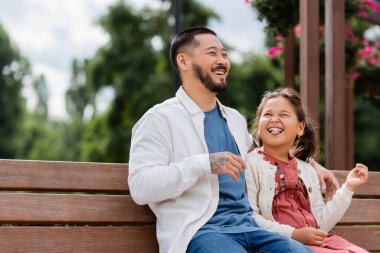 Asian kid sticking out tounge near cheerful dad on bench in park 