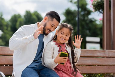Positive asian father and daughter having video call on smartphone on bench in park 