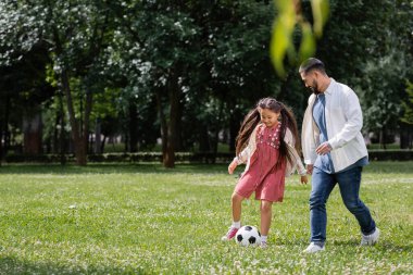 Cheerful asian kid playing football with dad on meadow in park 