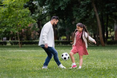Positive asian kid and parent playing football on meadow in park 
