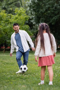 Cheerful asian dad playing football with blurred daughter in park 