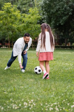 Happy asian father playing soccer with daughter on grass in park 