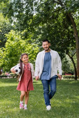 Smiling asian parent and child with soccer ball walking in park 