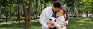 Cheerful asian dad holding football and hugging daughter in park, banner 