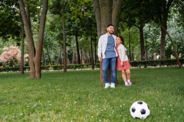 Asian father hugging preteen daughter near tree and soccer ball on grass 