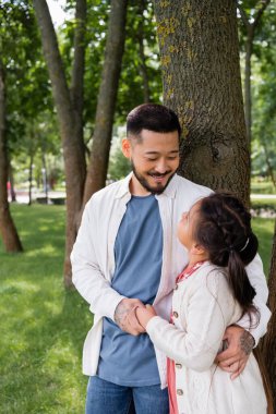 Smiling asian man holding hand and hugging preteen kid near tree in park 