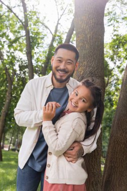 Positive asian father and daughter hugging and looking at camera in park 