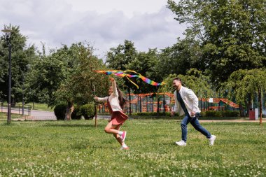 Cheerful asian girl playing with flying kite near dad in park 