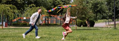 Happy asian girl holding flying kite while running near dad in park, banner 