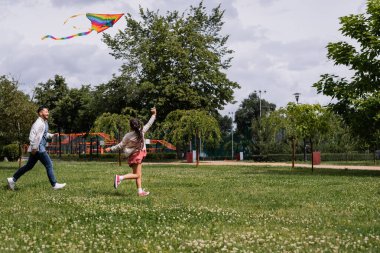 Preteen child holding flying kite near asian dad in park