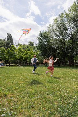 Positive asian parent running near daughter with flying kite in park 