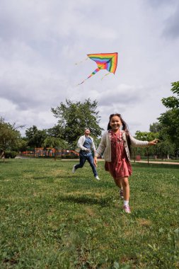 Happy asian girl running with flying kite near parent on lawn in park 