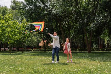 Smiling asian man holding colorful flying kite near daughter in park 