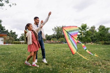 Smiling asian parent and kid looking at flying kite on lawn in park 