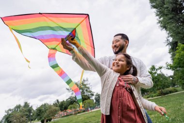 Positive asian dad hugging daughter with flying kite in summer park 