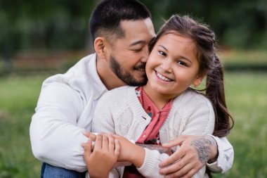 Portrait of asian dad hugging daughter looking at camera in park 