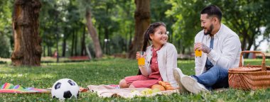 Asian child holding orange juice near food and dad in summer park, banner 