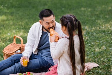 Child holding croissant near asian dad with orange juice in park 