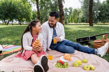Asian family holding orange juice and croissant during picnic in park 