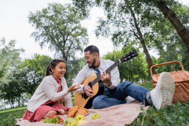 Smiling asian kid pointing with finger near father playing acoustic guitar near food in park 
