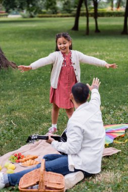 Positive asian girl looking at dad holding acoustic guitar near food on blanket in park 